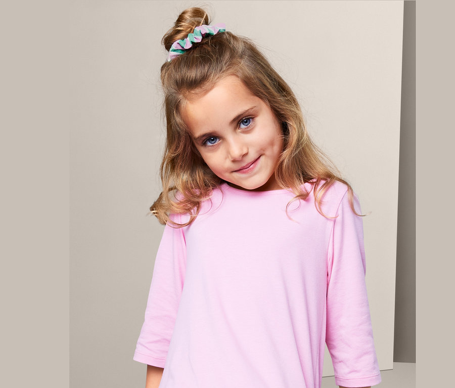 Ein blondes Mädchen mit blauen Augen trägt 3 Haargummis »Scrunchies« im Haar und ein rosa Kleid.