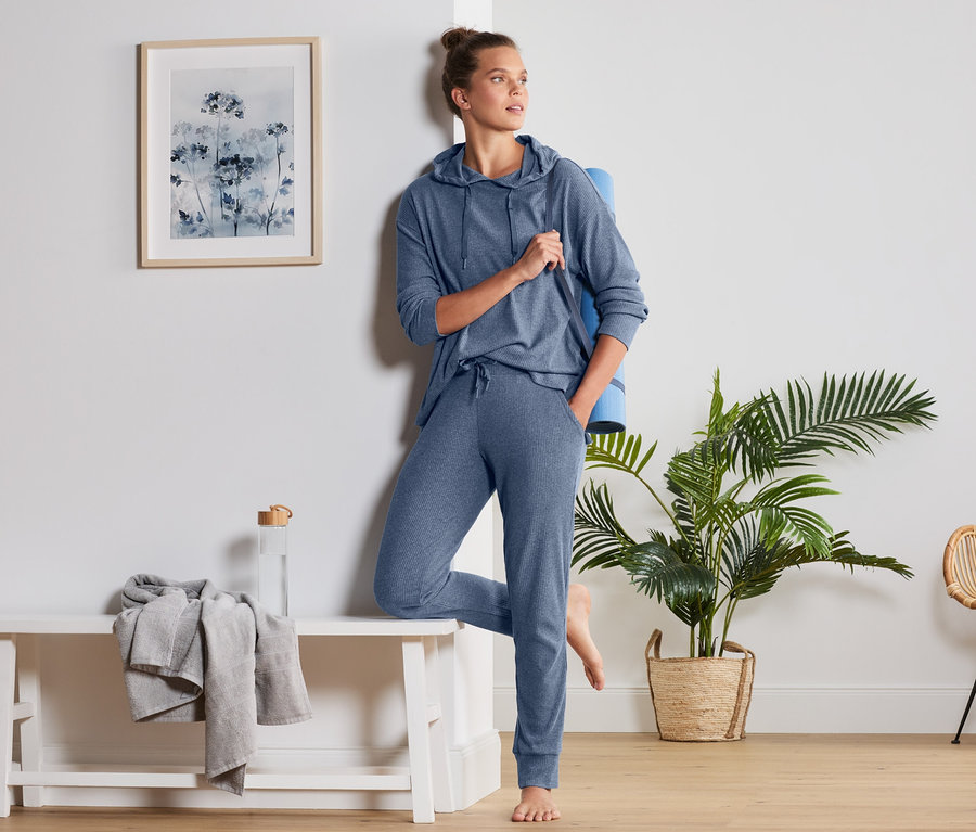Frau lehnt an der Wand, trägt blaue Relaxhose und Kapuzenpullover, Yogamatte unter dem Arm.