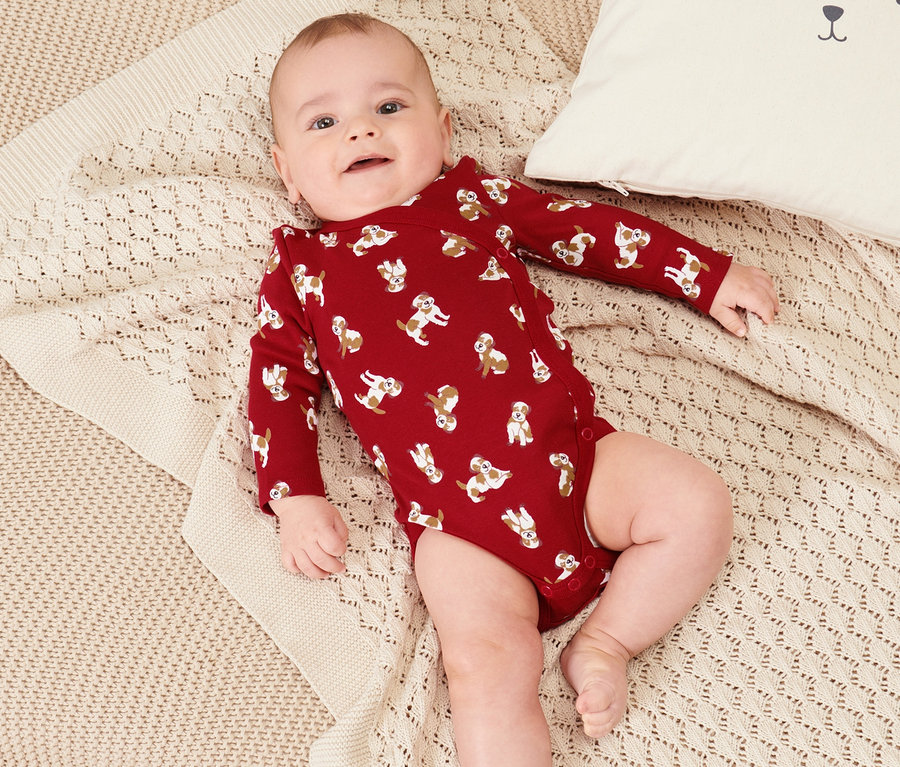 Baby liegt in einem 2er Set Baby-Bodys auf einer beigen Strickdecke neben einem weißen Kissen mit Bärengesicht.