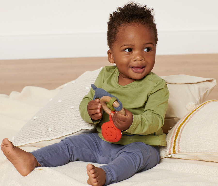 Lächelndes Baby sitzt auf einer Decke und hält ein Greifspielzeug.