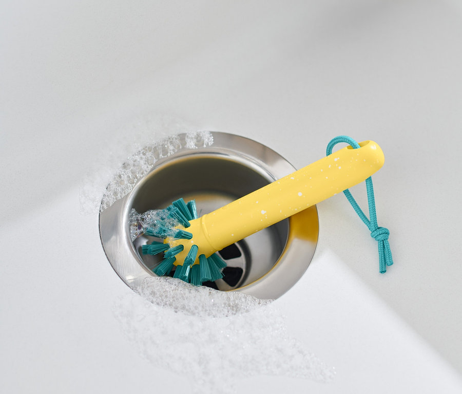 Gelbe Universalreinigungsbürste in Edelstahlspüle mit Schaum und beigem Handtuch.
