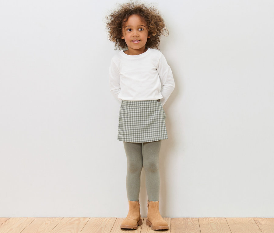 Mädchen steht und trägt einen Kinder-Hosenrock, ein weißes Oberteil und braune Schuhe.