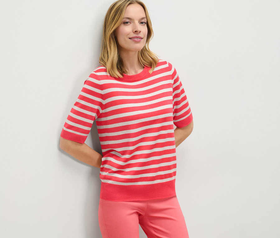 Frau lehnt an Wand, trägt gestreiften Feinstrickpullover und pinke Hose.