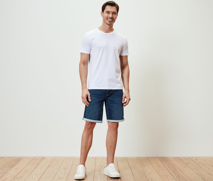 Mann steht und trägt Comfort Jeans-Shorts, ein weißes T-Shirt und Turnschuhe.