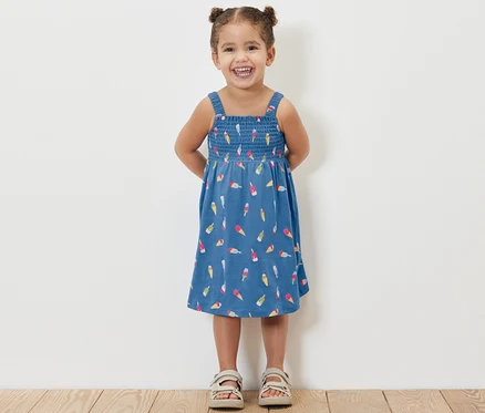 Ein lächelndes kleines Mädchen posiert in einem hellblauen Trägerkleid mit Eis am Stiel-Muster.