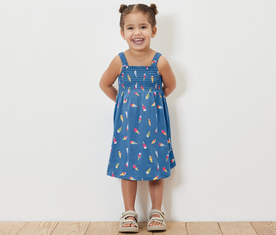 Ein lächelndes kleines Mädchen posiert in einem hellblauen Trägerkleid mit Eis am Stiel-Muster.