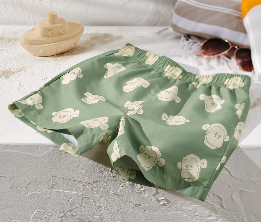 Grüne Kinder-Boardshorts mit Affenmuster liegen auf einer weißen Oberfläche neben einem Spielzeugboot, einem Handtuch, einer Sonnenbrille und Sonnencreme.