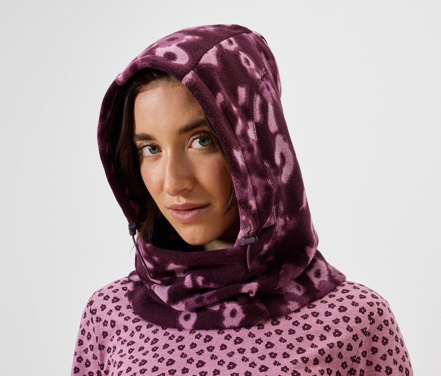 Porträt einer Frau mit braunen Haaren, die ein rosa Oberteil mit Muster und eine lila Schalmaske trägt.