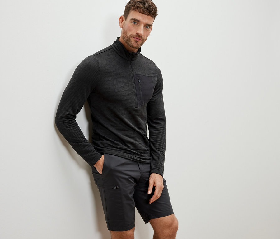 Mann lehnt an Wand, trägt anthrazitfarbenes Outdoor-Funktionsshirt und Shorts.