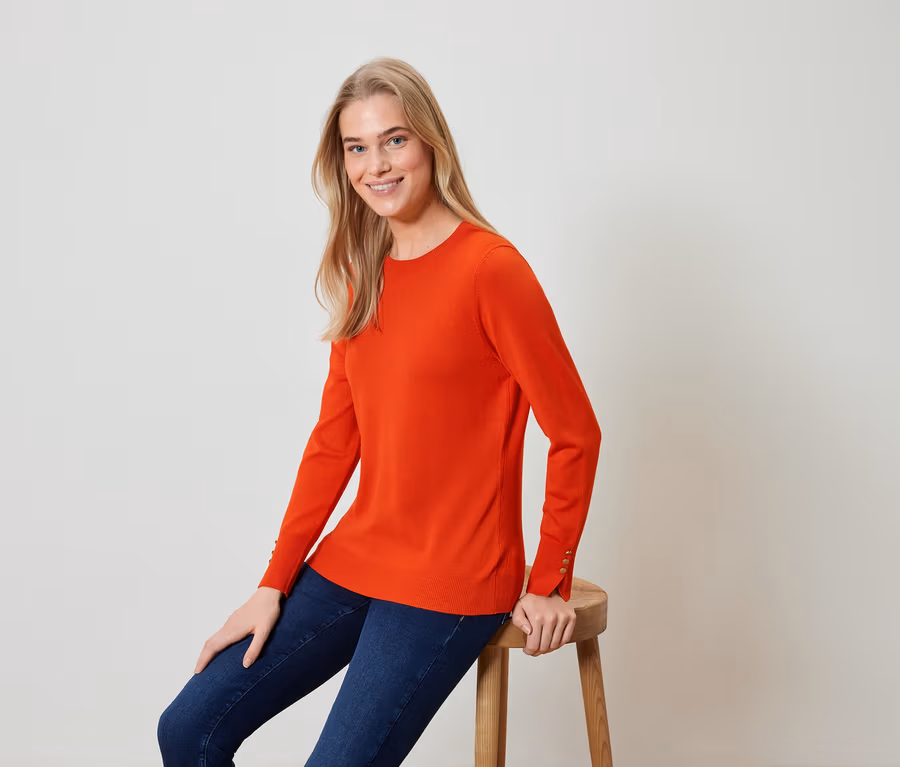 Blonde Frau sitzt auf einem Hocker und trägt einen orangefarbenen Pullover und blaue Jeans.