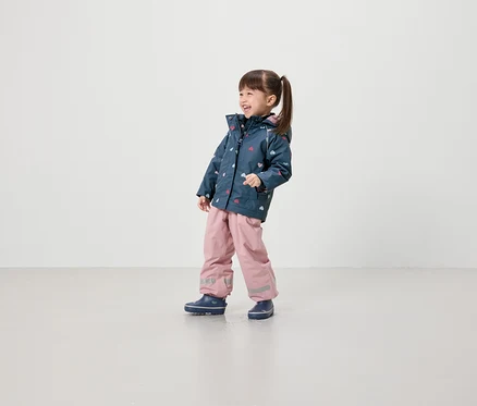 Ein lächelndes Mädchen in einer Jacke mit Herzmuster, rosafarbener Hose und blauen Schuhen steht vor einem hellen Hintergrund.