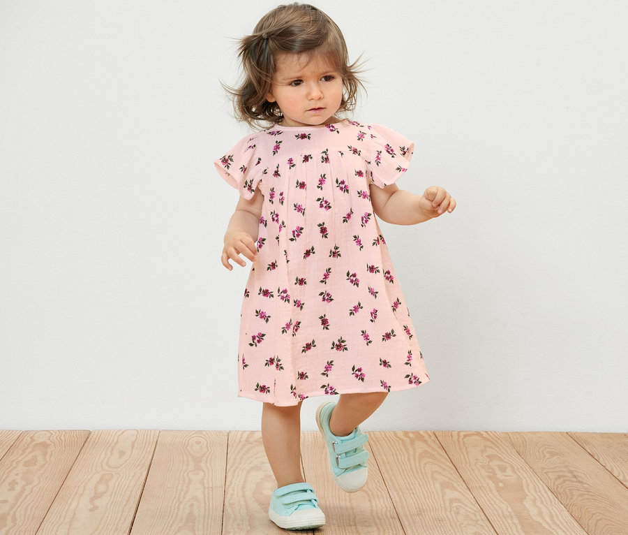 Ein kleines Mädchen in einem Baby-Musselinkleid mit Blumenmuster und blauen Schuhen steht auf einem Holzboden.