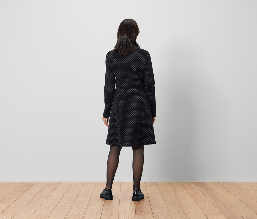 Eine Frau steht mit dem Rücken zur Kamera und trägt ein schwarzes Kleid und Strumpfhosen.