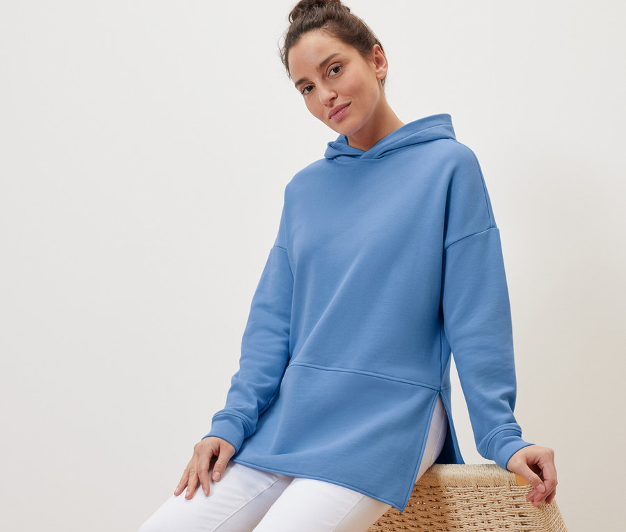 Frau sitzt auf einem Korbsessel und trägt ein hellblaues, langes Kapuzensweatshirt.