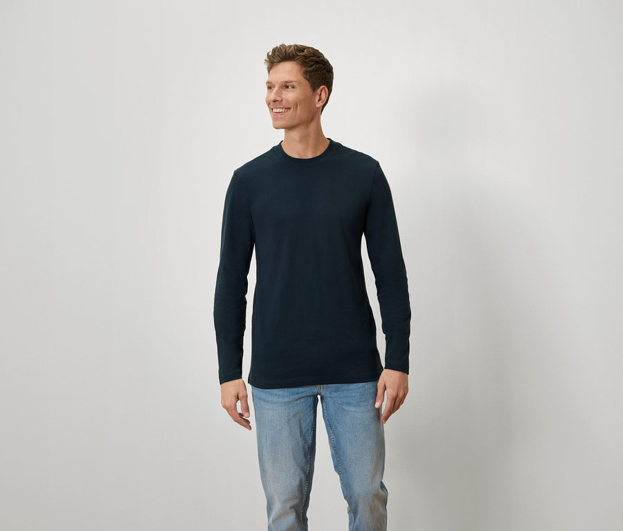 Mann mit braunen Haaren und blauen Augen trägt ein blaues Langarmshirt und Jeans.