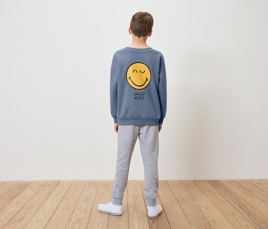 Junge steht mit dem Rücken zur Kamera und trägt ein blaues Sweatshirt mit gelbem Smiley-Motiv und graue Trainingshose.