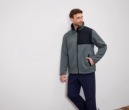 Ein Mann lehnt an einer Wand und trägt eine graue Fleecejacke mit schwarzen Schultern und dunkelblaue Hosen.