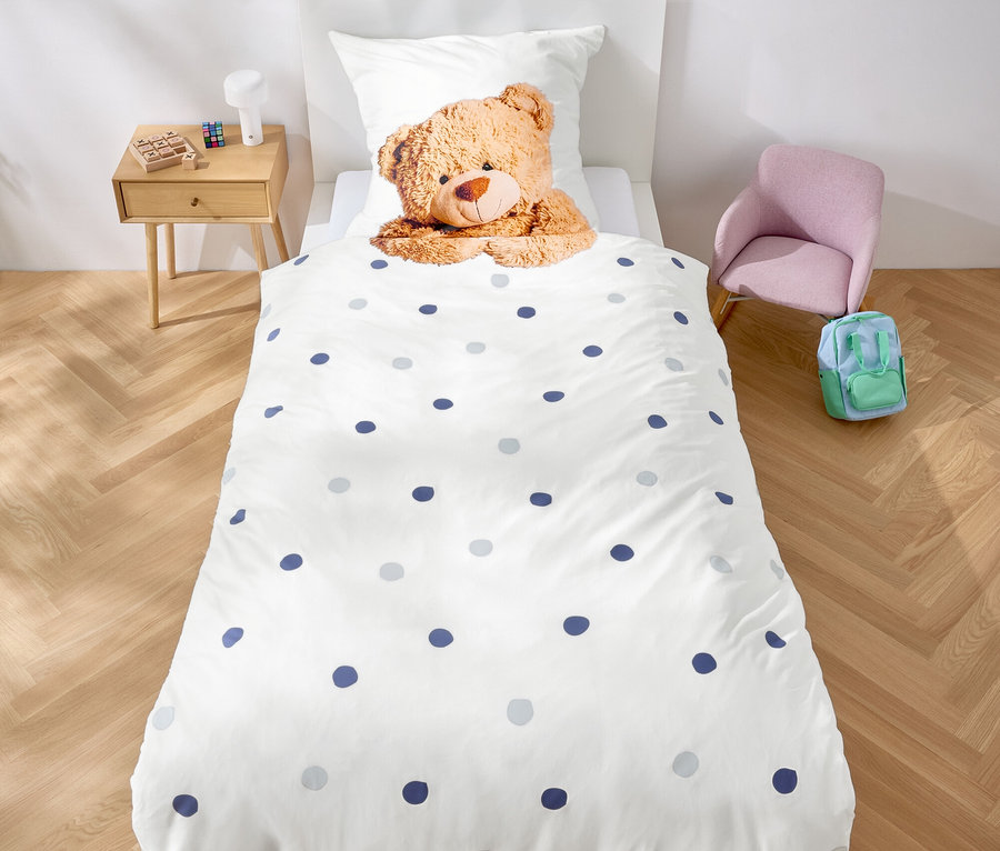 Im Zimmer steht ein Bett mit weißer Bettwäsche mit blauen Punkten und einem Teddybär. Über dem Bett hängt eine Girlande aus lila Dreiecken.
