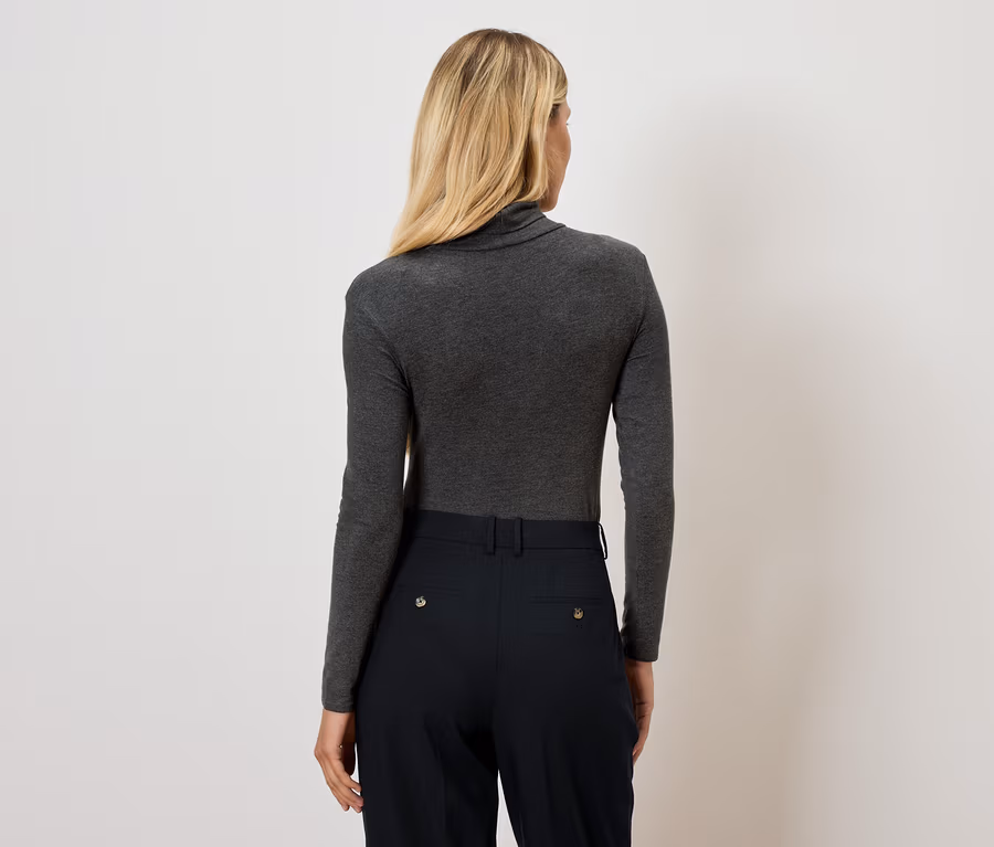 Rückansicht einer Frau mit blonden Haaren in einem grauen Rollkragenpullover und einer schwarzen Hose.