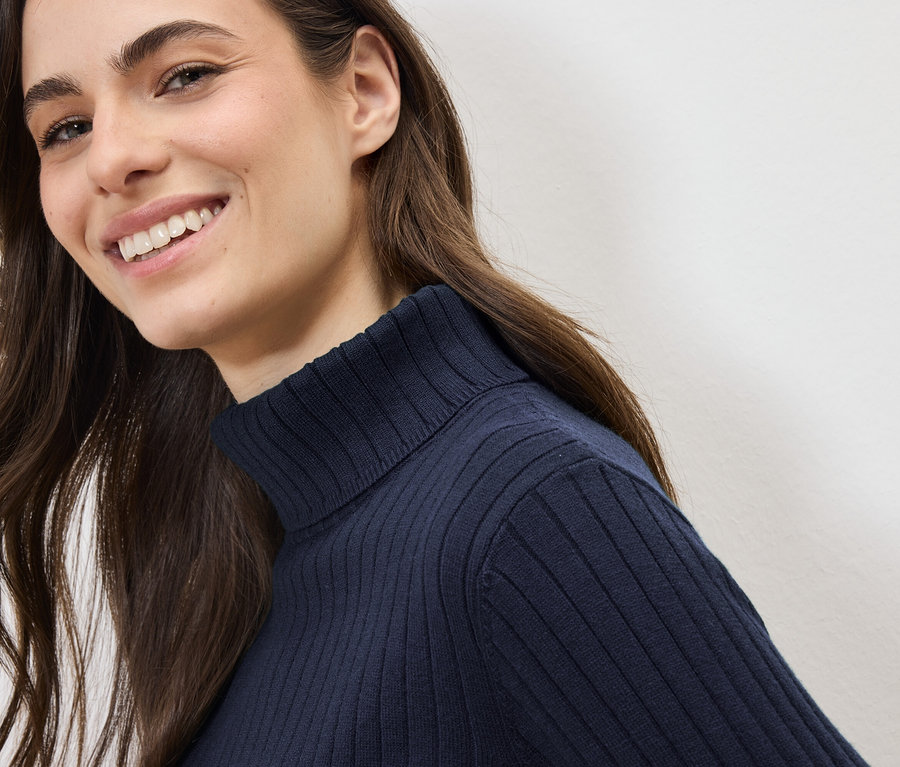 Lächelnde Frau mit langen braunen Haaren trägt einen dunkelblauen Rollkragenpullover.