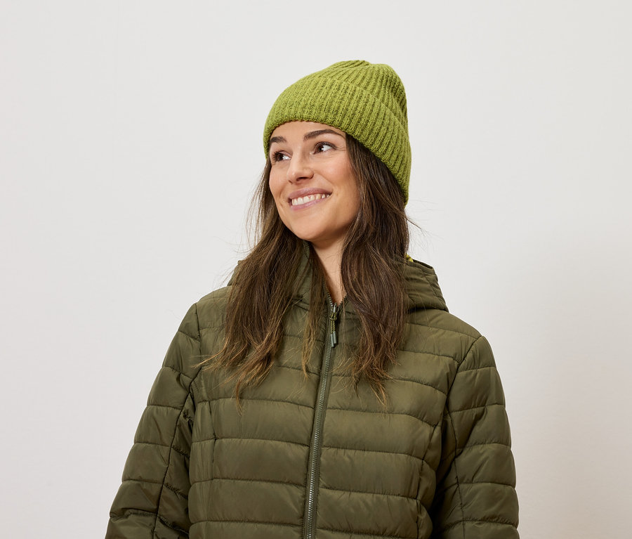 Lächelnde Frau trägt eine olivgrüne Steppjacke und eine Strickmütze.