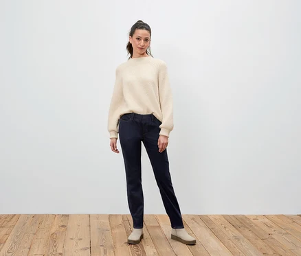 Frau steht und posiert in einem cremefarbenen Pullover, dunkelblauen Hosen und beigen Schuhen.
