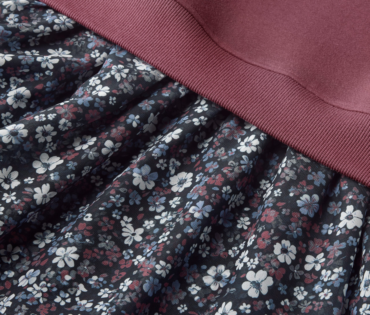 Detailansicht eines Kinder-Sweatkleids mit Blumenmuster auf dem Rockteil und bordeauxfarbenem Oberteil.