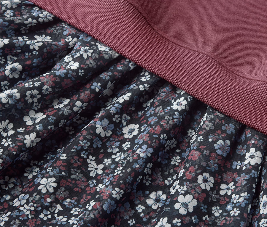 Detailansicht eines Kinder-Sweatkleids mit Blumenmuster auf dem Rockteil und bordeauxfarbenem Oberteil.