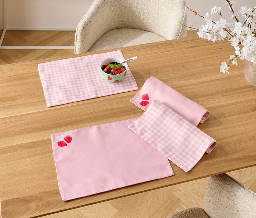 Auf einem Holztisch liegen rosa Platzsets, eines mit Erdbeermuster, eine Schale mit Beeren und eine Vase mit blühenden Zweigen.
