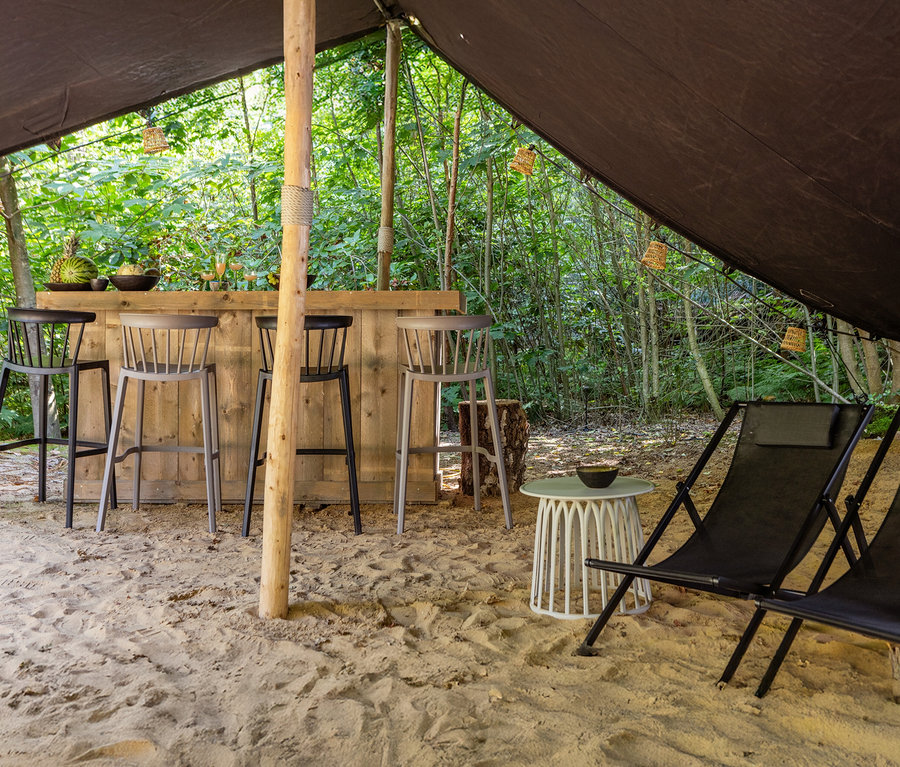 Barhocker stehen vor einer Holzbar in einem Zelt im Wald. Liegestühle und ein Beistelltisch stehen im Sand.