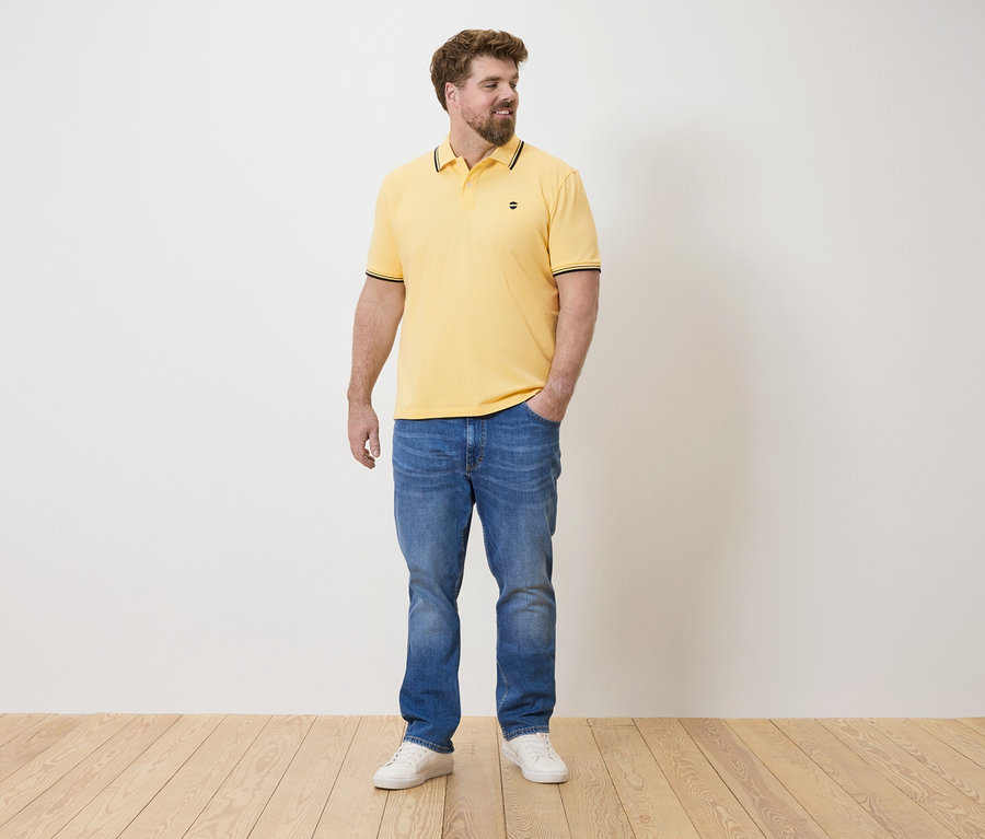 Ein Mann steht mit einer Hand in der Hosentasche, trägt ein gelbes Poloshirt, blaue Jeans und weiße Turnschuhe.