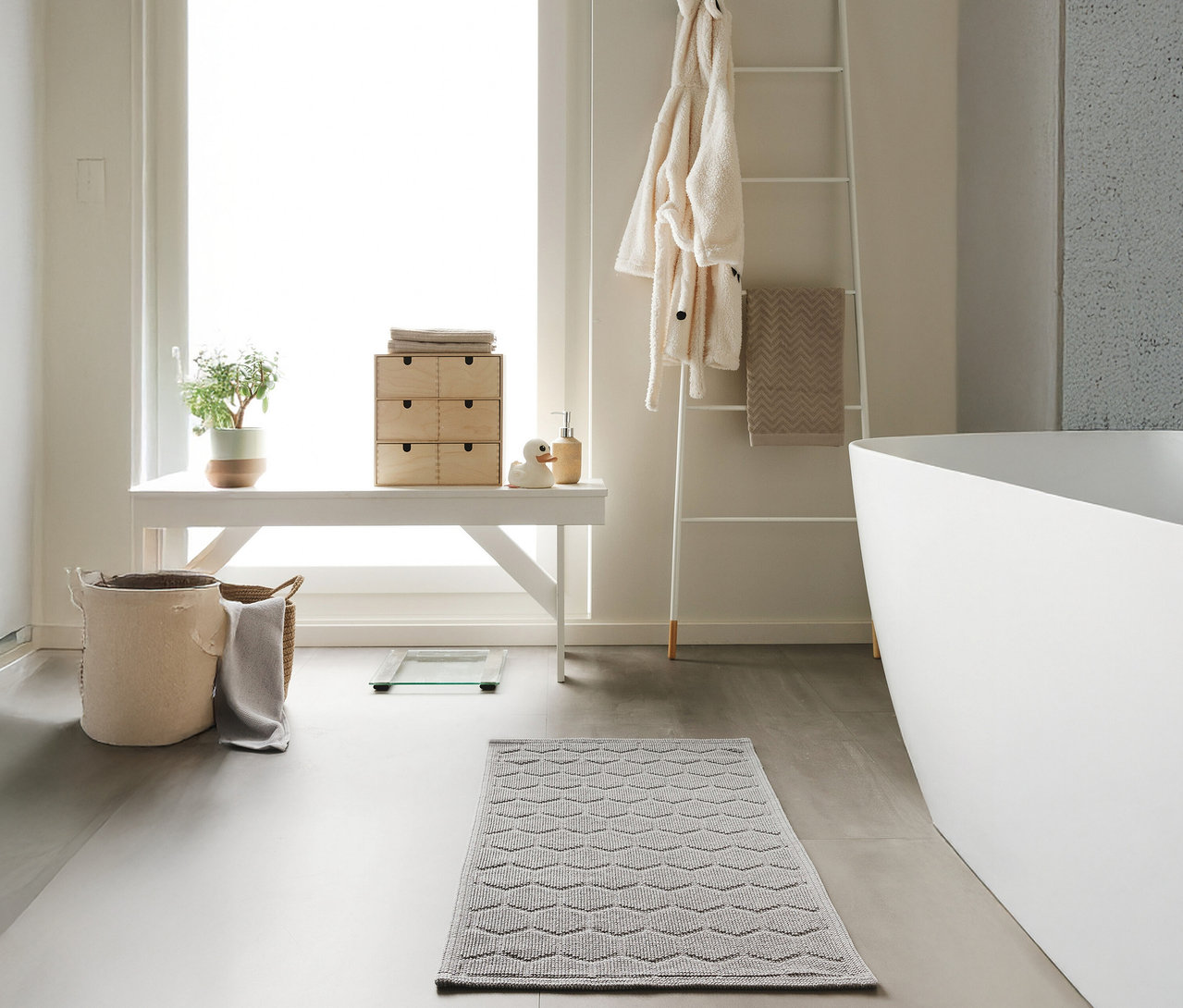 Ein Badezimmer mit einer weißen Bank, einer Leiter und einer Badewanne. Auf dem Boden liegt eine hellgraue Badematte »Jojo« von benuta mit Rautenmuster (50 x 80 cm).