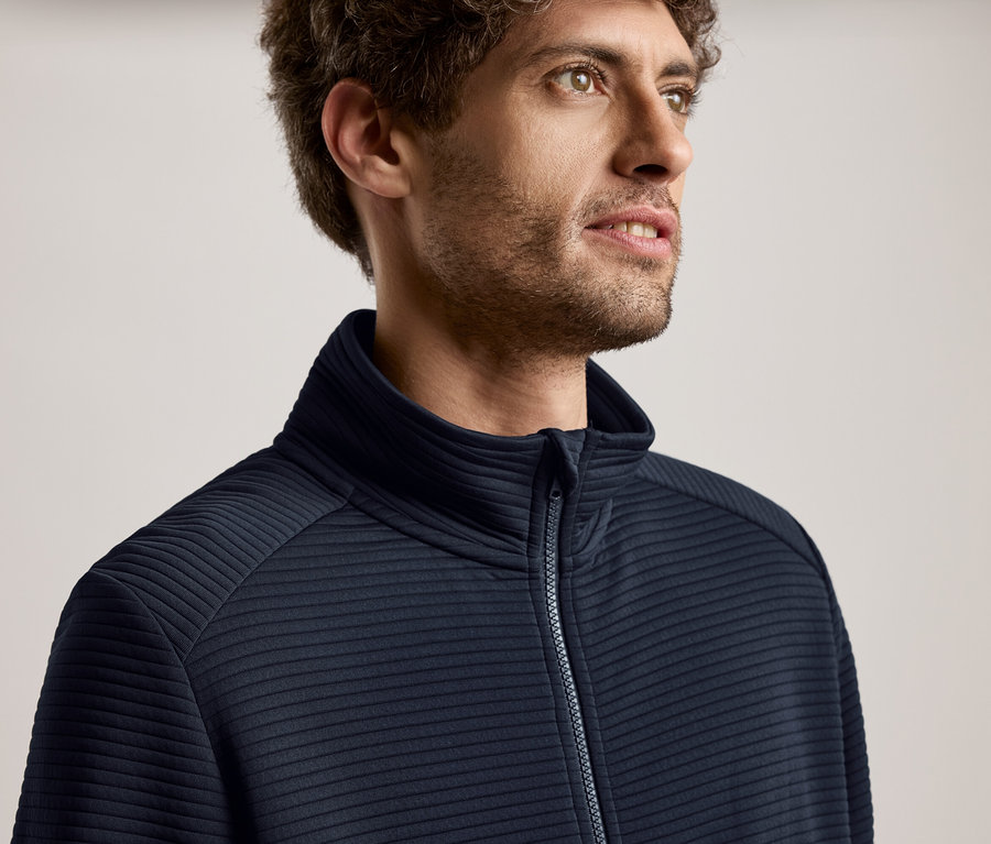 Nahaufnahme eines Mannes mit lockigem Haar, der eine dunkelblaue Jacke mit Reissverschluss trägt und nach rechts blickt.