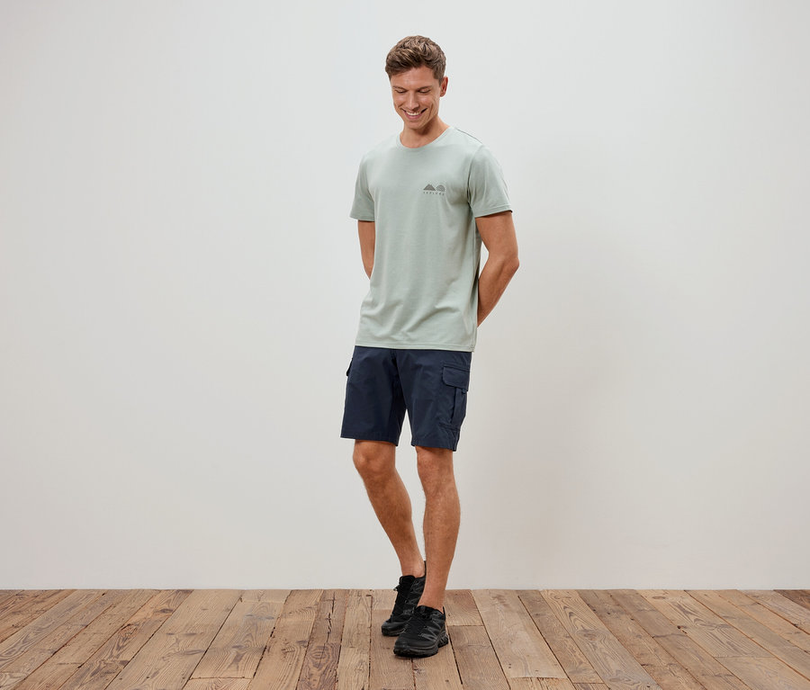 Mann steht vor einer Wand, trägt ein hellgrünes T-Shirt, blaue Shorts und schwarze Schuhe.