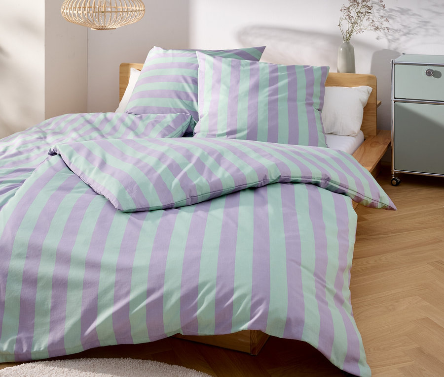 Ein Bett mit gestreifter Bettwäsche in Lila und Grün steht in einem hellen Raum.
