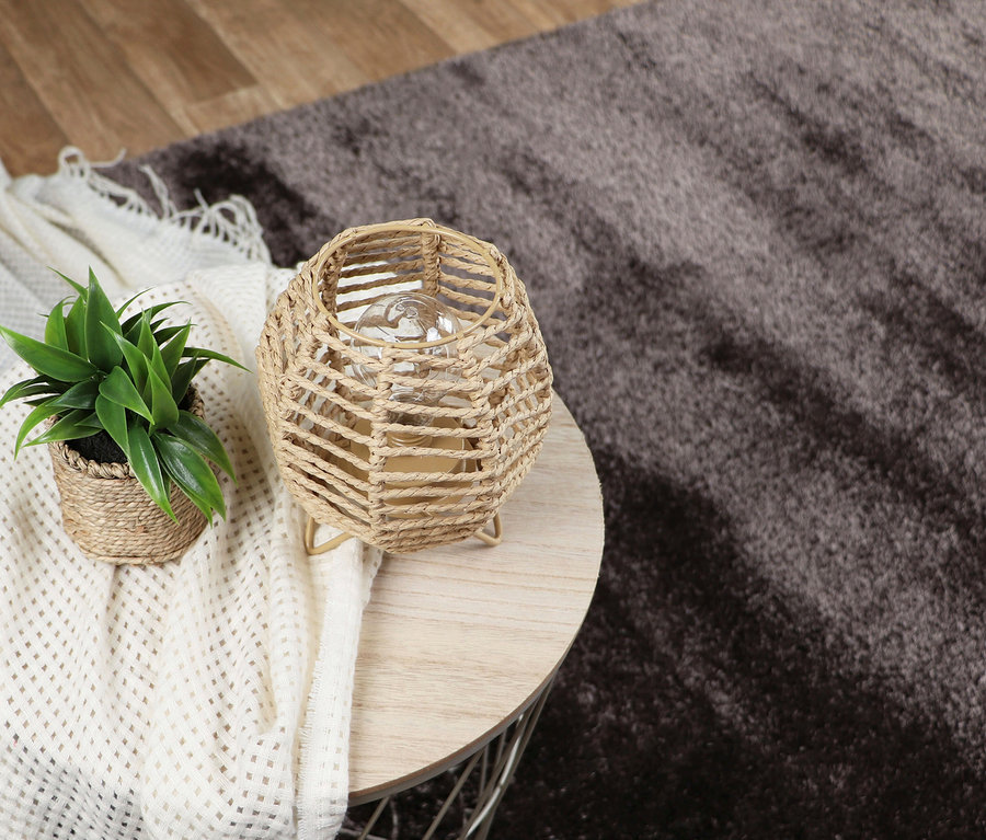Nahaufnahme eines runden Tisches mit einer Pflanze, einer Lampe und einer weißen Decke, im Hintergrund ein dunkelbrauner Teppich »Silky«.