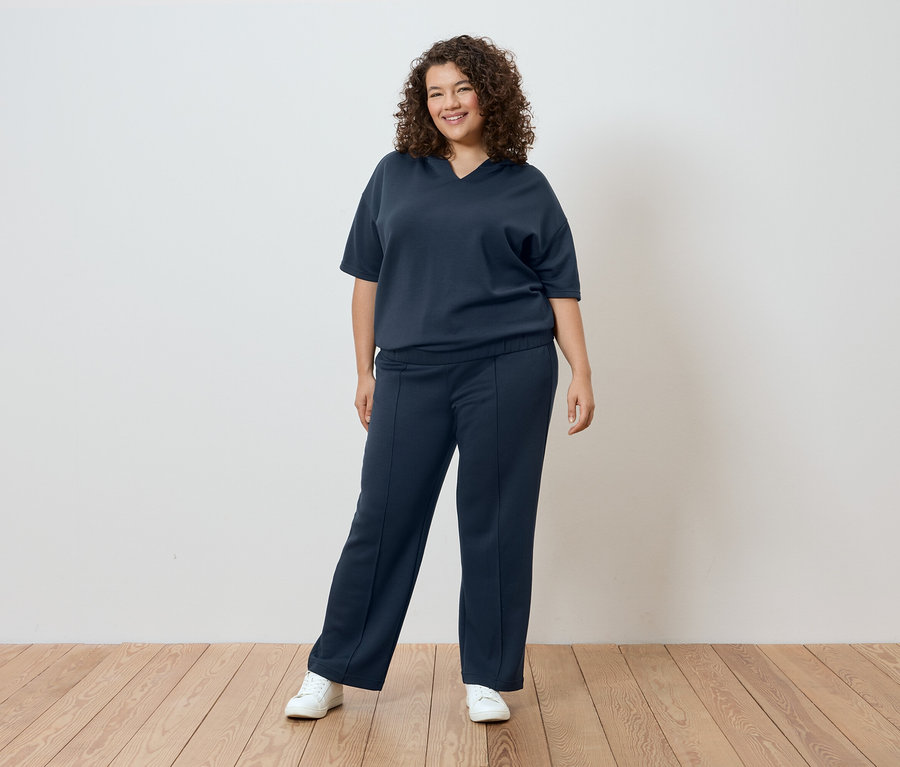 Frau mit lockigem Haar trägt ein dunkelblaues T-Shirt und eine Hose vor einer weißen Wand.