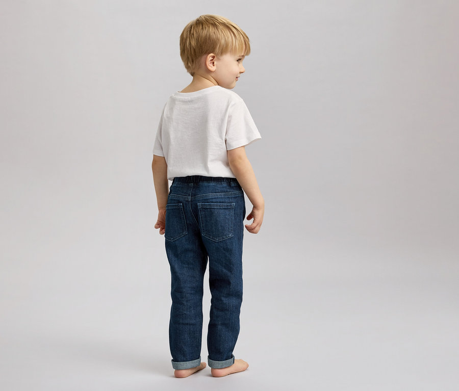 Ein blonder Junge mit weißem T-Shirt und Jeans steht barfuß vor einem grauen Hintergrund und schaut nach rechts.