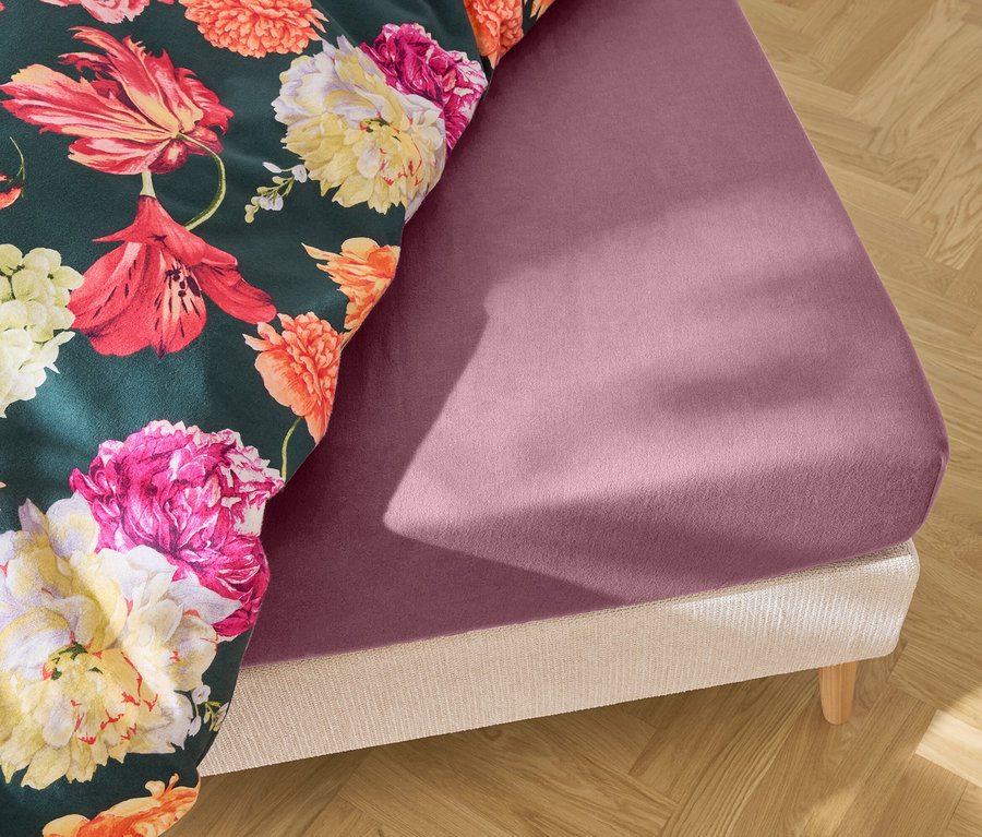 Nahaufnahme eines Bettes mit einem lila Spannbettlaken und einer Blumendecke auf einem Holzboden.