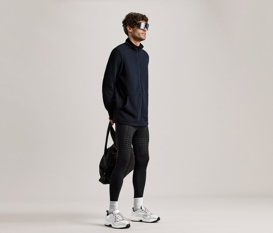 Mann mit Sonnenbrille, schwarzer Jacke, Leggings und Turnschuhen hält eine schwarze Tasche.