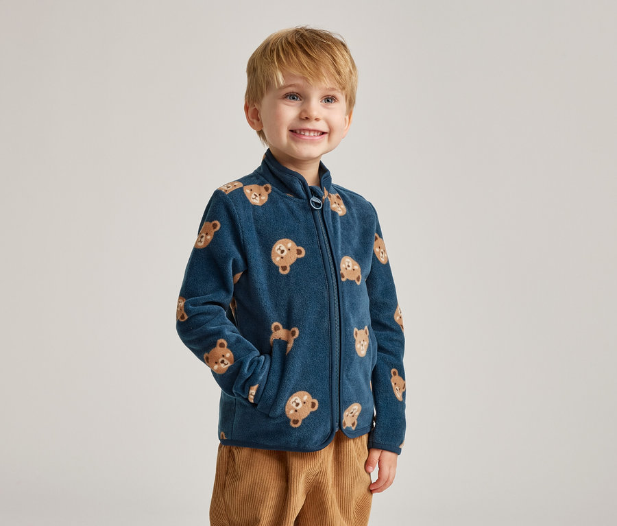 Ein lächelnder Junge mit blonden Haaren trägt eine marineblaue Fleecejacke mit Bärenmotiven und braune Cordhosen.