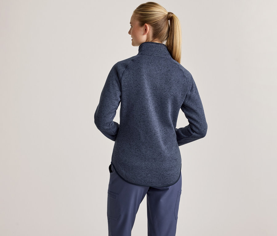 Eine Frau von hinten, mit blondem Pferdeschwanz, trägt eine blau gesprenkelte Fleecejacke mit Stehkragen und blaue Hosen.