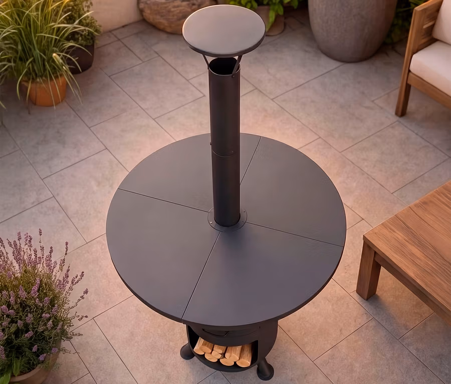 Dunkelgraue, runde Outdoor-Feuerstelle mit hohem zentralem Schornstein, kleiner runder Ablagefläche und Holzablage am Sockel auf einer gepflasterten Terrasse.