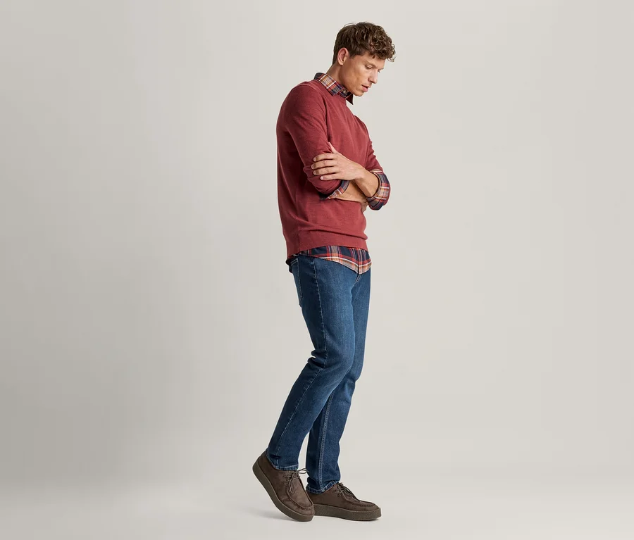 Ein Mann in einem roten Pullover über einem karierten Hemd, blauen Jeans und braunen Schuhen, der mit verschränkten Armen nach unten blickt.
