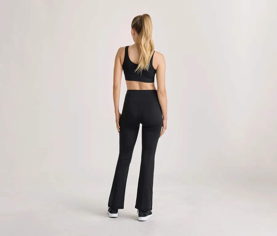 Frau steht mit dem Rücken zum Betrachter, trägt einen schwarzen Sport-BH, schwarze High-Waist-Leggings mit Schlag und schwarze Turnschuhe, die blonden Haare zum Pferdeschwanz gebunden.