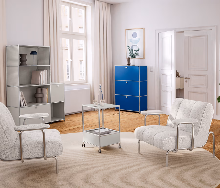 Helles Wohnzimmer mit Holzboden und weißer Einrichtung. Links ein hellgraues Regal, in der Mitte zwei helle Sessel und ein Beistelltisch auf einem runden Teppich. Rechts ein blauer Schrank und eine Pflanze.