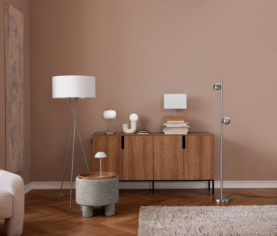 Ein modernes Wohnzimmer mit hellbraunen Wänden und Holzboden. Links ein cremefarbener Sessel. Eine Stehlampe mit Stativ steht neben einem grauen Hocker mit kleiner Lampe. Ein Holzsideboard trägt zwei Tischlampen, ein Dekoobjekt und Bücher. Rechts eine silberne Stehlampe mit zwei Kugelleuchten, ein heller Teppich liegt auf dem Boden.