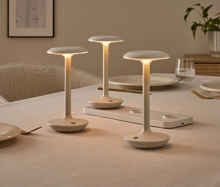 Drei minimalistische, cremefarbene Tischlampen auf einem gedeckten Esstisch, eine davon auf einer Ladestation. Die Lampen leuchten warm.