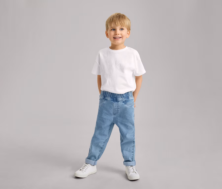 Ein lächelnder kleiner Junge steht in einem weißen T-Shirt, hellblauen Kinder-Pull-on-Pants – Fit »Luca« mit hochgekrempelten Hosenbeinen und weißen Turnschuhen.