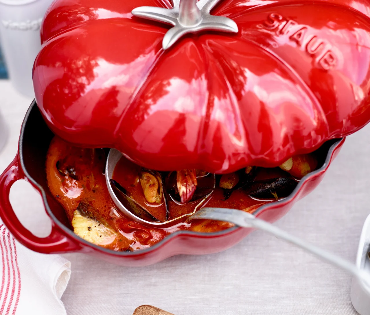 Roter STAUB Gusseisen-Bräter »Tomate« mit teilweise geöffnetem Deckel, gefüllt mit einem Eintopf und einer Schöpfkelle.